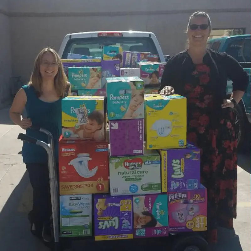 about webp2 Two women stand beside a cart loaded with various brands of diapers and baby supplies, in front of a pickup truck.